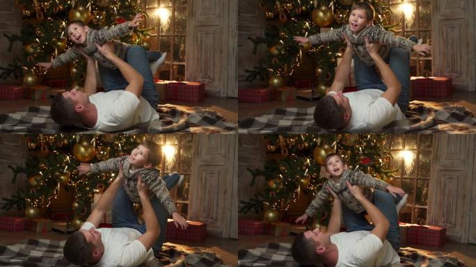 父亲躺在客厅温暖的木地板上，装饰着一棵发光的圣诞树，抱起他的小儿子，享受节日快乐的概念