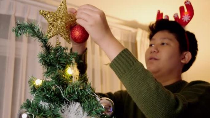 快乐的亚洲男孩戴着驯鹿发带，晚上在家装饰圣诞树，节日庆祝概念。