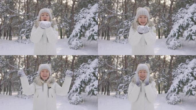 快乐可爱的女孩，穿着温暖的冬衣，把手中的雪吹向相机，然后把它抛起来。美丽模糊的背景积雪的自然在松树林