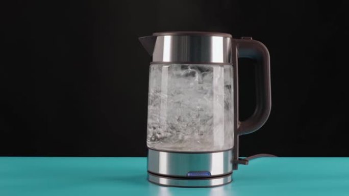 现代玻璃电热水壶，放在蓝色桌子上，黑色背景，装满水烧开。在运动中沸腾。用来制造饮料和开水。
