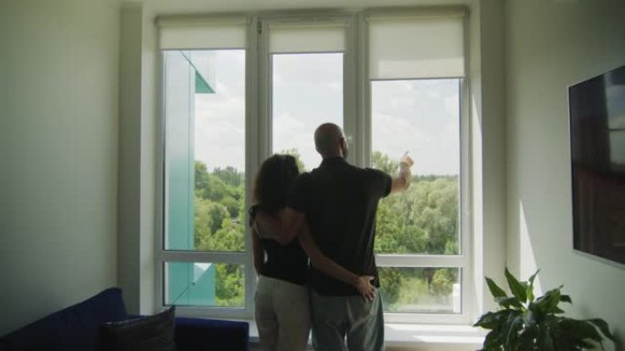 站在窗前的年轻夫妇的后视图。男人在外面展示一些东西