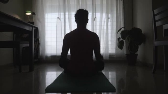一个年轻人坐在瑜伽垫上的剪影运动镜头，在客厅里以冥想或盘腿的姿势。