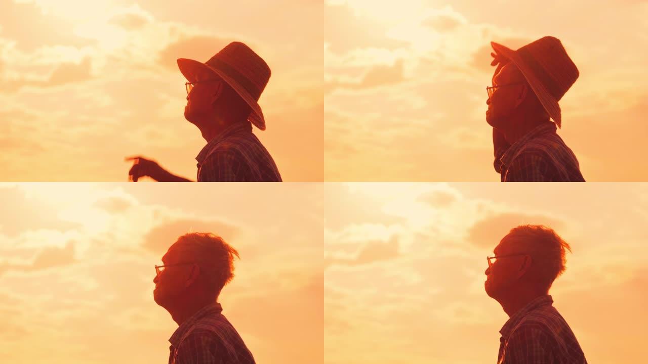 日落时站在稻田里的高级农民的剪影。一位戴着帽子的老人在金色的天空背景上向远处望去。