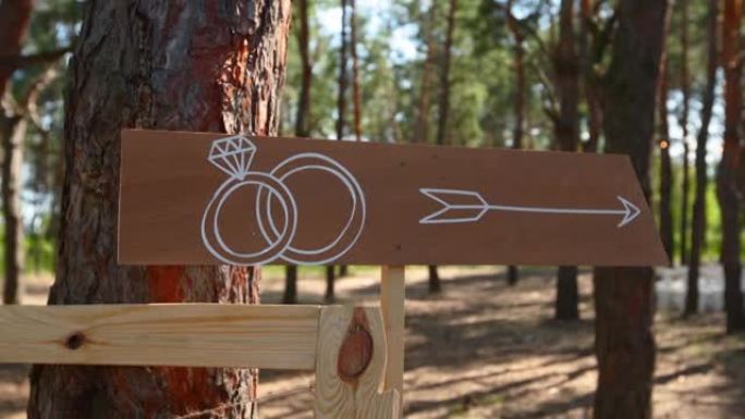 仪式场地上有结婚戒指的木制箭头标志。婚礼派对横幅帖子。户外松树林客人的方向信息横幅。乡村风格或乡村风