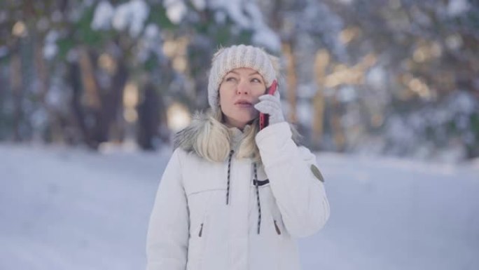 穿着温暖的冬装可爱的女孩的肖像为手机聊天并欢欣鼓舞。年轻女子在白雪皑皑的森林模糊背景下在户外摆姿势。