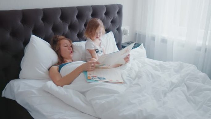 晨床阅读。年轻女子在明亮的现代卧室的床上为她蹒跚学步的孩子读这本书