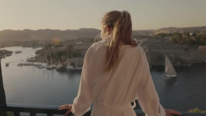 女人站在阳台上，在日落时欣赏壮丽的河景。日落时漂浮在尼罗河上的帆船。