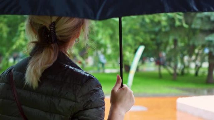 一位金发女子在雨中撑着雨伞站在城市公园，雨滴浸湿了她的外套。概念: 自然、水、全球变暖