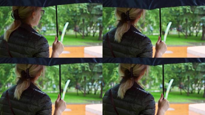 一位金发女子在雨中撑着雨伞站在城市公园，雨滴浸湿了她的外套。概念: 自然、水、全球变暖