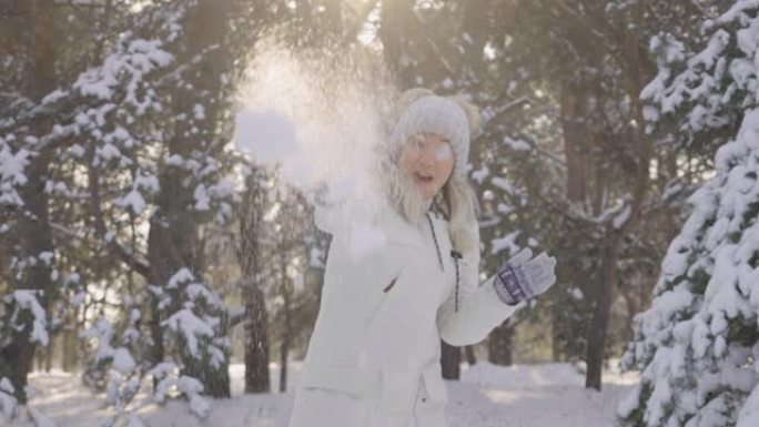 快乐可爱的女孩穿着冬装，对着镜头扔雪球，开心地笑着。下雪的冬天，阳光灿烂。美丽的模糊背景雪域自然松林