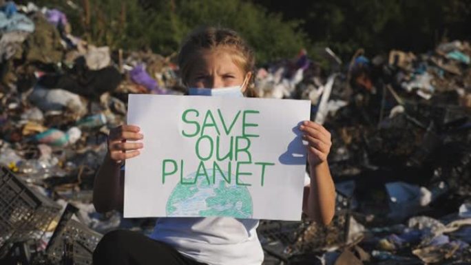 戴着医用防护面具的小女孩看着相机，手持环保运动海报。小生态活动家在垃圾的背景下举起拯救自然的标语牌。