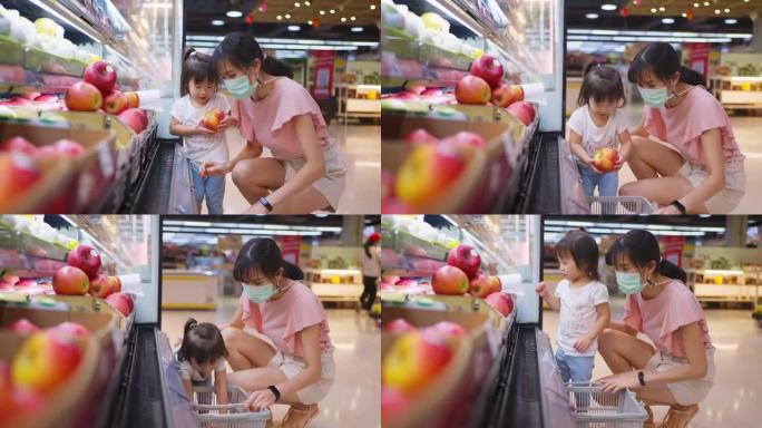 年轻的亚洲美丽的母亲拿着菜篮子，带着孩子在超市散步，戴着口罩，以防止COVID感染。她正在选择红苹果