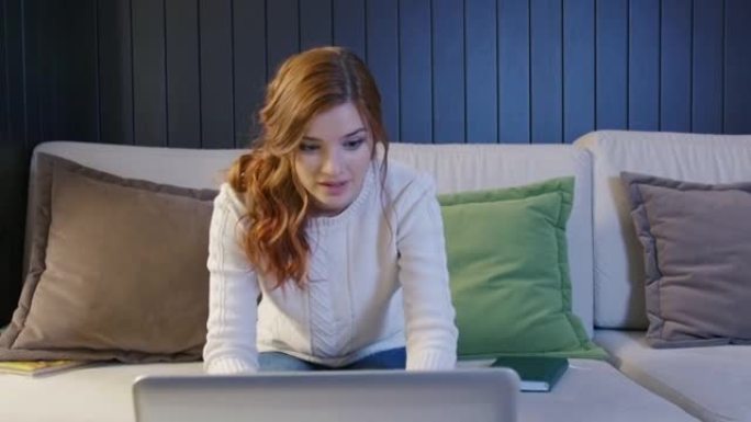 兴奋的年轻女性获胜者看着笔记本电脑庆祝在线成功坐在家里的沙发上。欣快的女士获得了新的远程工作机会