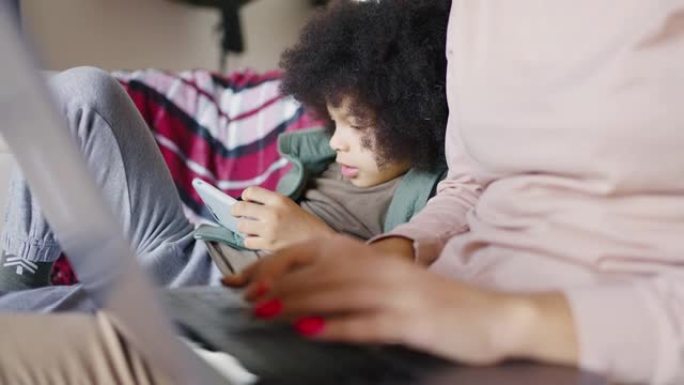 母亲在笔记本电脑上工作的手和坐在笔记本电脑旁边的孩子使用智能手机的特写镜头。混血男孩在妈妈工作时打电