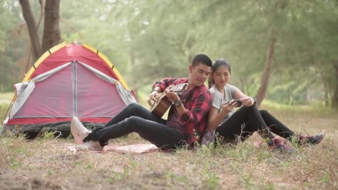 徒步旅行和露营概念。一对亚洲夫妇在树林里露营时一边弹吉他一边看手机一边唱歌。