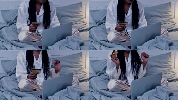 漂亮的非洲裔美国妇女使用便携式笔记本电脑和信用卡在线购物。黑人女性穿着睡衣在卧室的床上摆姿势。家庭房