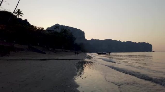 泰国甲米镇美丽的海滩是在日落或日落时拍摄的，长尾船回到海滩的前面，而太阳落在背景山脉上。
