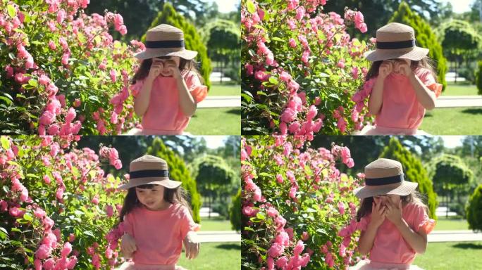 五岁的白人女孩，一头棕色长发，穿着粉红色的t恤和草帽，在阳光明媚的日子里，在公园的玫瑰丛附近用手揉眼