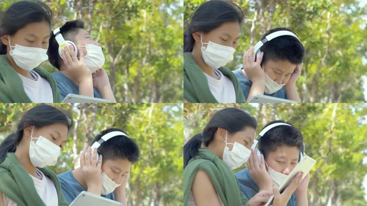 快乐的亚洲学童戴着口罩，在校园户外用数字平板电脑和耳机放松，社交疏远的生活方式理念。