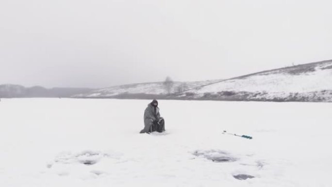带着钓鱼竿的渔夫正坐在冰冻池塘的冰洞附近。人在冬季钓鱼时抓鱼，空中无人机视图。