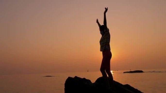 女孩的剪影站在岩石上，在海滩上举手，早晨日出，生活方式的概念。