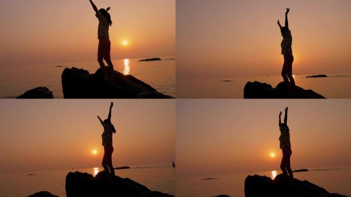 女孩的剪影站在岩石上，在海滩上举手，早晨日出，生活方式的概念。