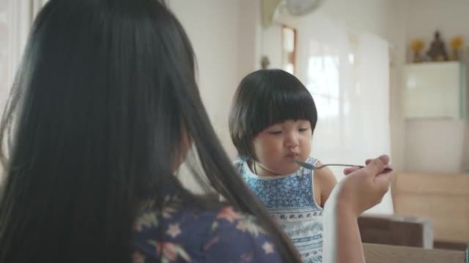 年轻的母亲在餐桌上用汤匙喂她的小女儿吃饭。可爱的亚洲幼童女孩与她的母亲吃午餐。