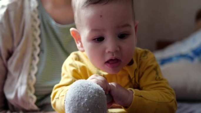 一位年轻的母亲抱着一个小孩坐在床上，用牙胶和其他触觉玩具控制游戏。母性、母性和家庭观念。慢动作