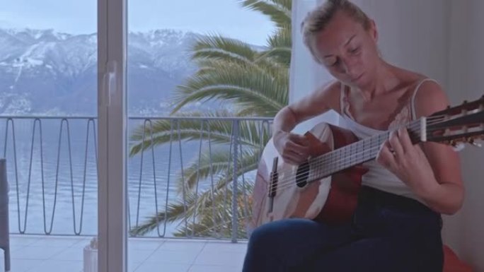 年轻女性在家弹吉他。窗外的湖泊和山脉景色