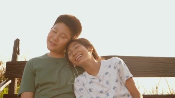 快乐的亚洲人和他的妹妹一起用耳塞听音乐，同时坐在户外公园的木制秋千上，带着早晨的阳光，生活方式的概念