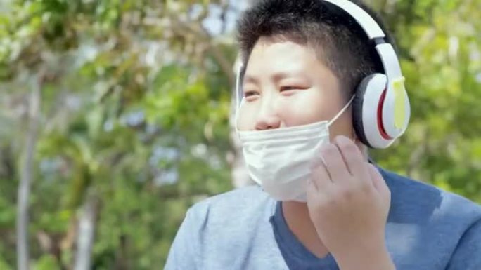 快乐的亚洲男孩戴着口罩，在户外校园里用无线耳机听音乐时摇头，社交距离概念。