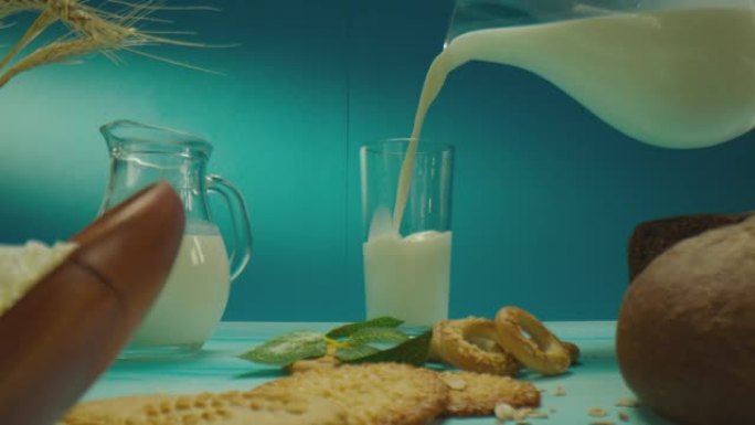 装饰桌子的美丽组成，带有牛奶，乳制品，烘焙产品，浆果，蓝色背景上的花朵。男人从水罐里把牛奶倒进玻璃杯