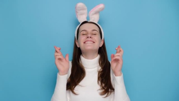 可爱的年轻高加索女性在交叉手指时许愿，表情积极，戴着兔子蓬松的耳朵和白色套头衫，模特在蓝色工作室背景
