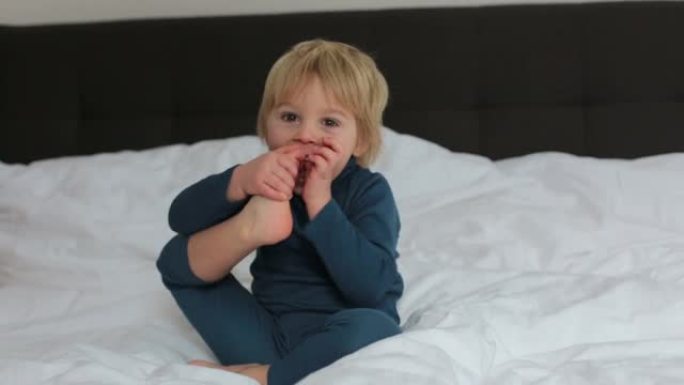 可爱的金发蹒跚学步的孩子，吮吸他的脚拇指，做有趣的脸，笑