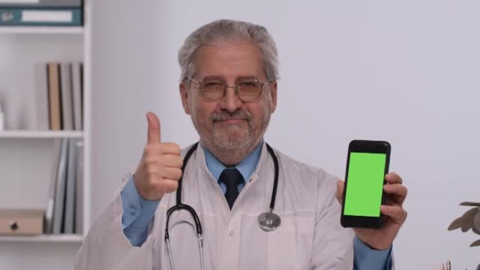 老年男性医生拿着带有绿屏色度键的智能手机，用手指指着它，并做拇指向上的手势。穿着白色医用外套的医生坐