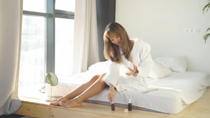 可爱的亚洲女人，洗完澡后坐在床上，为身体选择化妆品。阳光落在床上