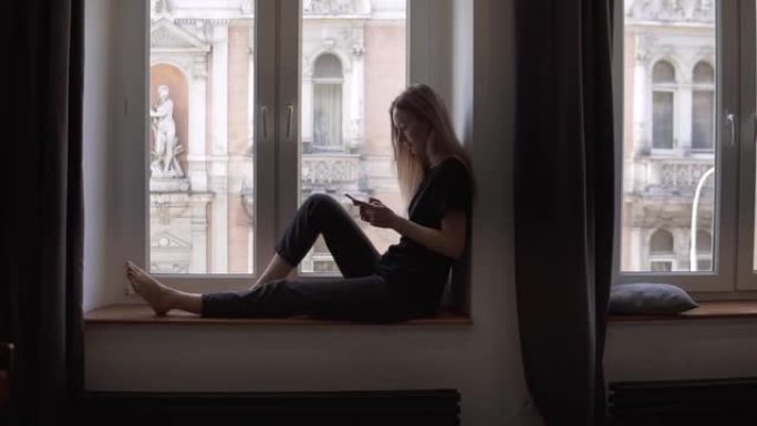 金发女人坐在窗台上，在客厅用智能手机和钢琴