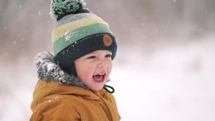 冬季降雪背景下快乐蹒跚学步男孩的肖像。穿着黄色派克大衣的婴儿欢喜第一场雪。慢动作。
