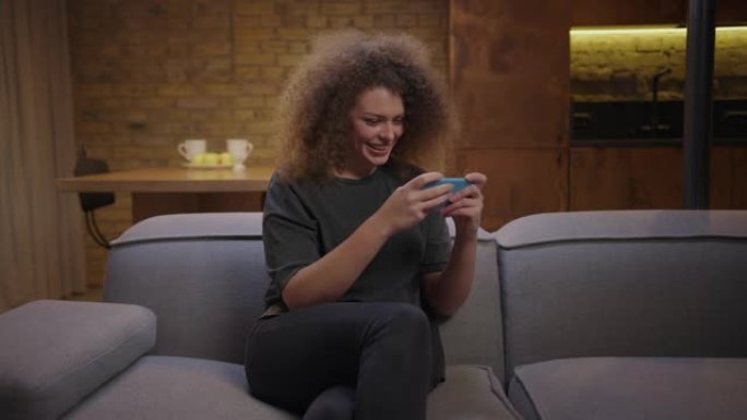 千禧一代的女人在手机上玩手机游戏，坐在沙发上赢得游戏。女性在玩手机时用手做胜利手势。