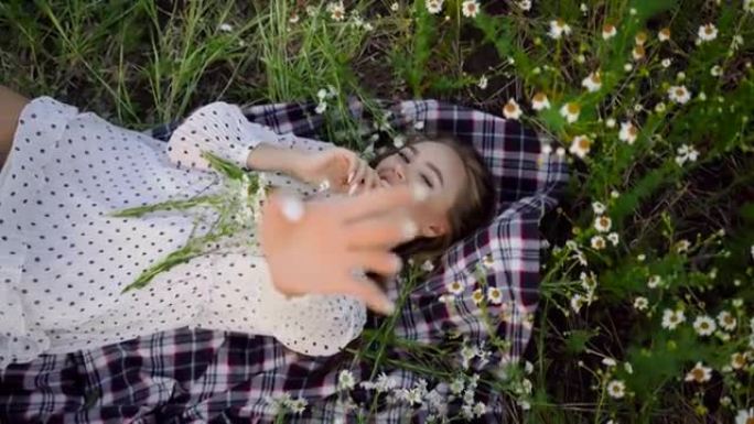 一位美丽的年轻女子躺在雏菊地里的格子布上，摆出姿势让摄影师拍照，看着相机