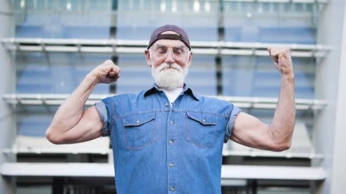 戴着棒球帽的强壮老人展示肌肉，在相机上眨眼，力量