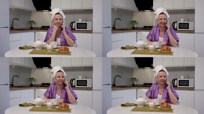 年轻女子坐在厨房的桌子旁，打电话，准备吃早餐和喝咖啡。穿着紫色浴袍和毛巾的女性在灯光室的背景下摆姿势