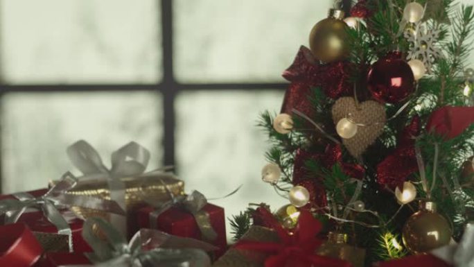 新年圣诞节背景，背景上有霜冻的窗户和飘落的雪。圣诞树，礼物。装饰品，红色的金球，发光的灯泡。温暖的家
