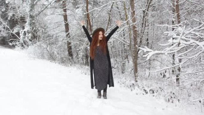 一个穿着黑色连衣裙和帽子的红色长发的年轻女孩站在白雪覆盖的森林里时伸了个懒腰