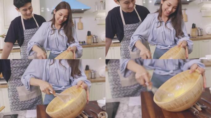 亚洲夫妇在家里的厨房里互相帮助，在浪漫的气氛中制作面包店。年轻人一起工作，将配料混合在木碗中，然后将