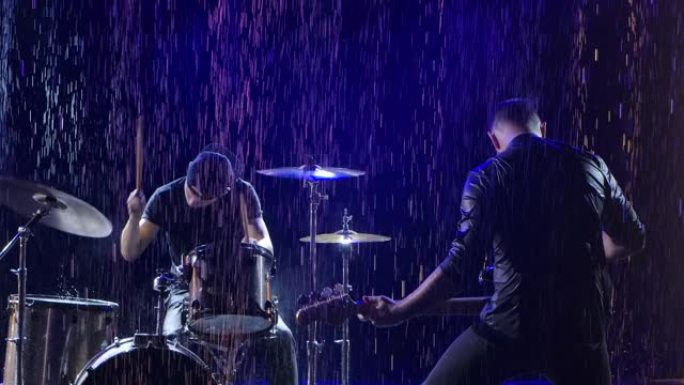 摇滚乐队在倾盆大雨中被蓝光照亮的黑暗工作室中的情感表演。鼓手和贝斯手正在现场举行摇滚音乐会。特写。慢