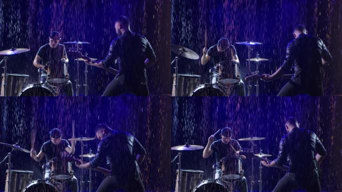 摇滚乐队在倾盆大雨中被蓝光照亮的黑暗工作室中的情感表演。鼓手和贝斯手正在现场举行摇滚音乐会。特写。慢