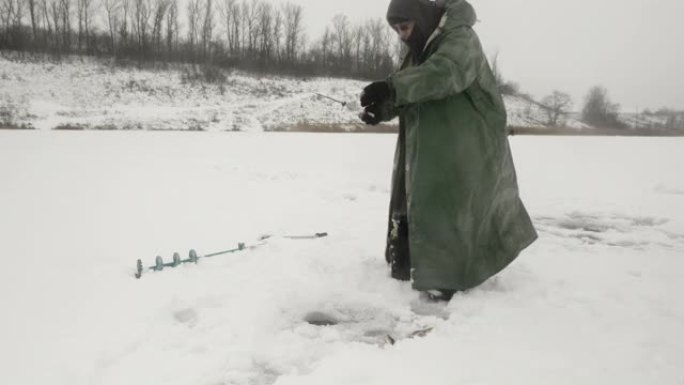 冬季钓鱼概念。穿着冬季极端衣服的渔夫正在冰洞里抓鱼。人在冰冻的湖上钓鱼