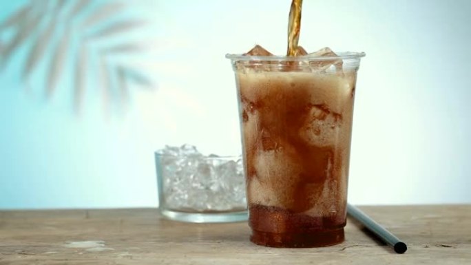 新鲜的可乐饮料在塑料外卖杯中与冰块形成泡沫和飞溅，背景中散焦的棕榈叶。慢动作