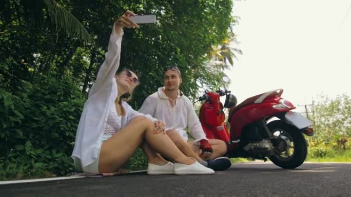 两个白人旅游女人坐在红色滑板车附近。让它自拍。穿着白色衣服骑摩托车的情侣在森林之路小径旅行。